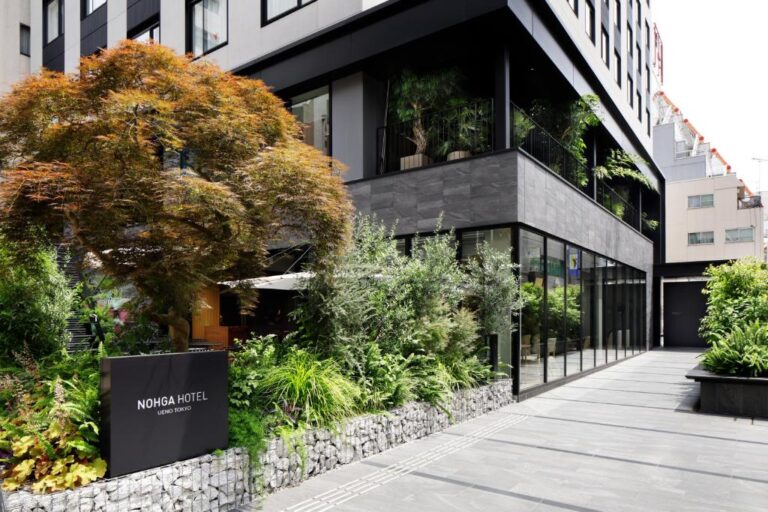 上野諾嘉飯店 (NOHGA HOTEL UENO TOKYO) 充滿自然綠意的入口外觀，以現代洗鍊的深色建築材質搭配豐富植栽，在熱鬧的上野商圈中打造出一座極具質感的靜謐綠洲。